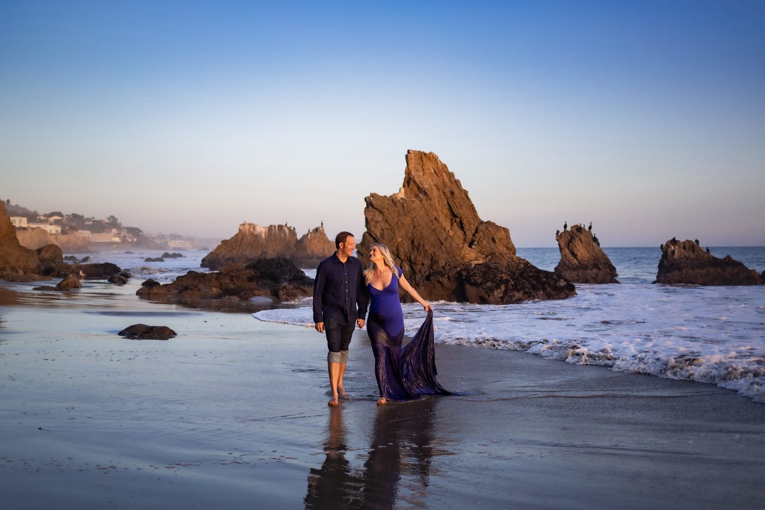 A couple walking hand in hand along the shoreline at sunset, she in a flowing blue gown