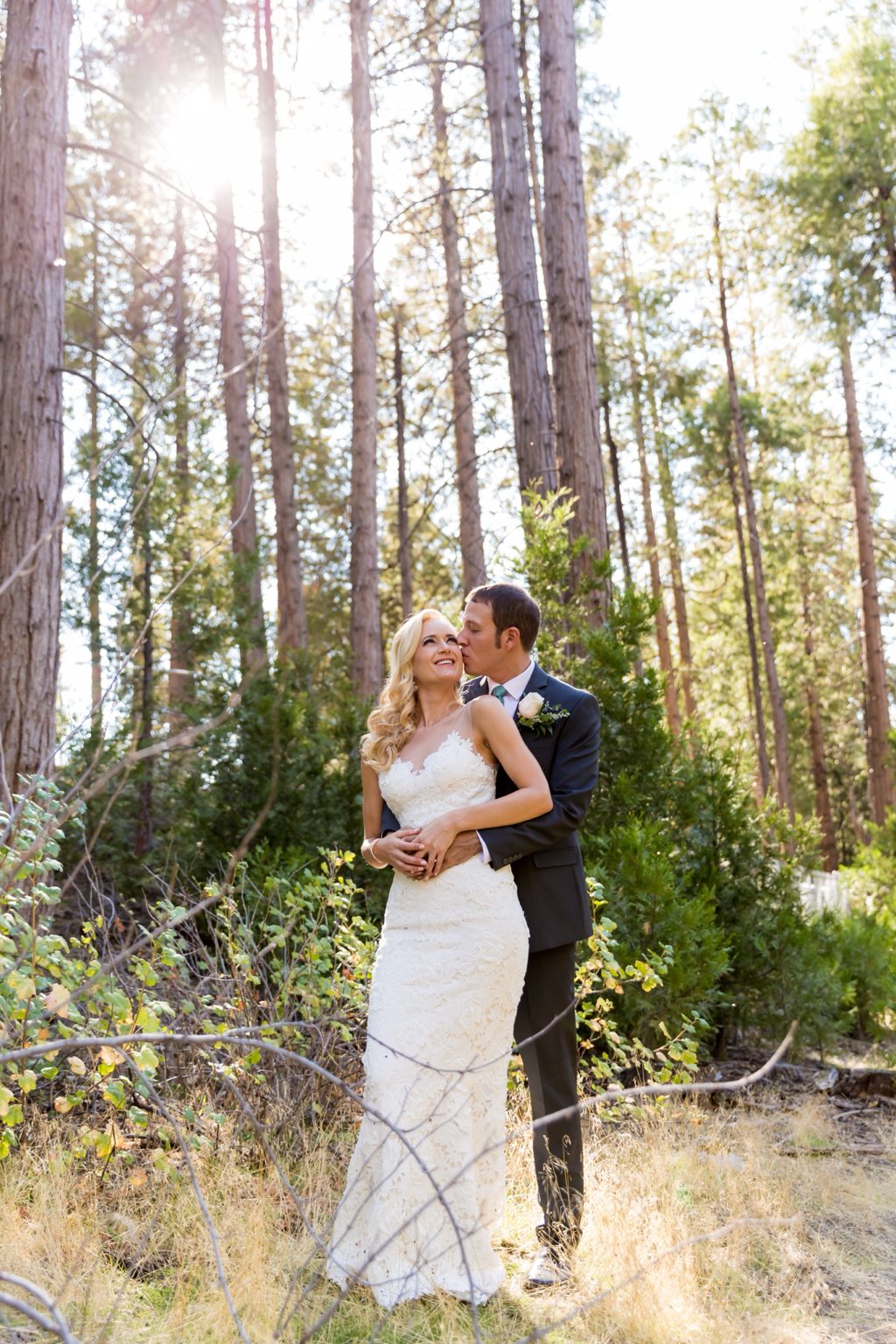 A groom holding his bride from behind in a sunlit forest, kissing her temple