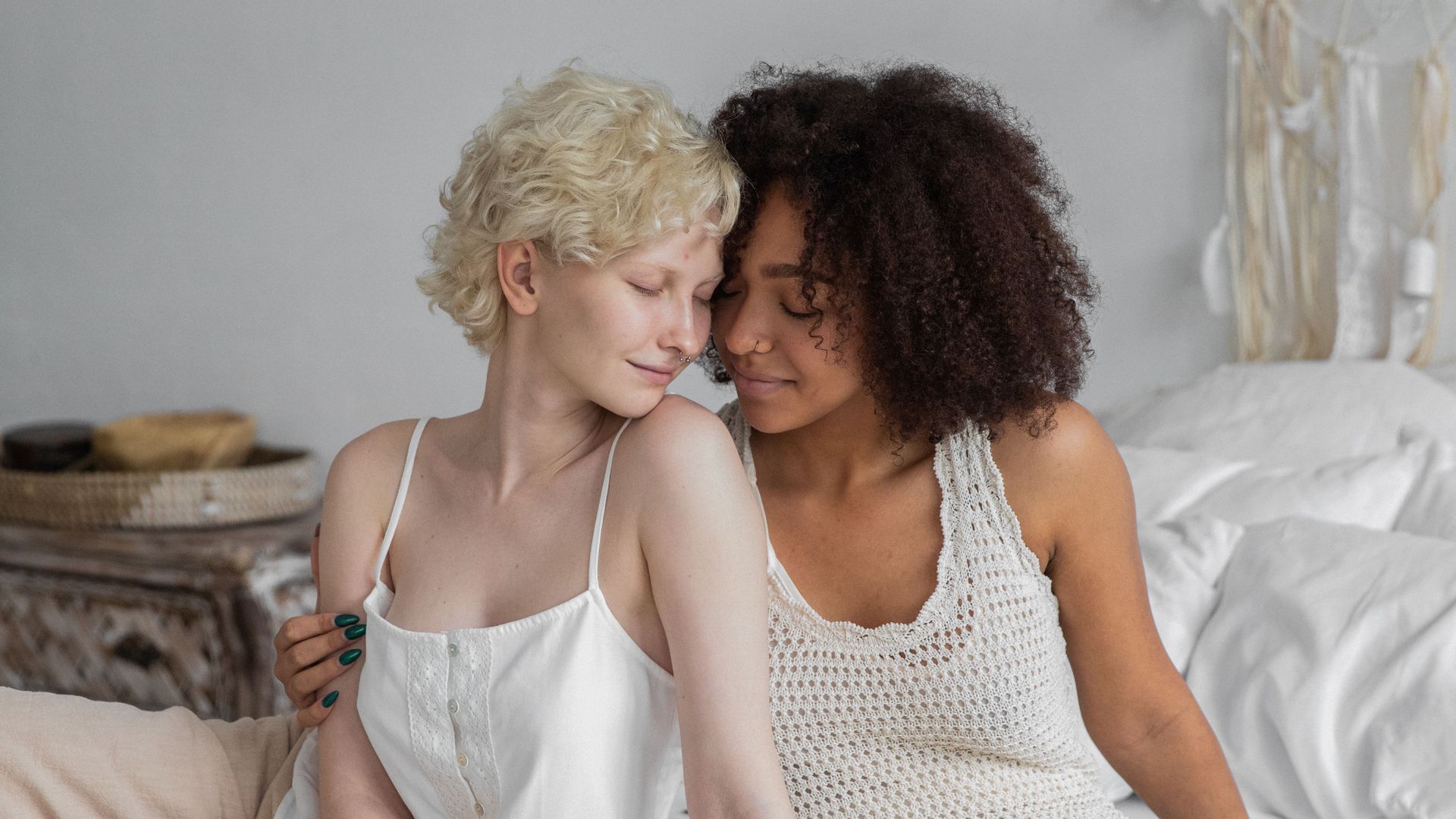 Two women resting forehead to forehead in a soft, sunlit bedroom, eyes closed in tenderness