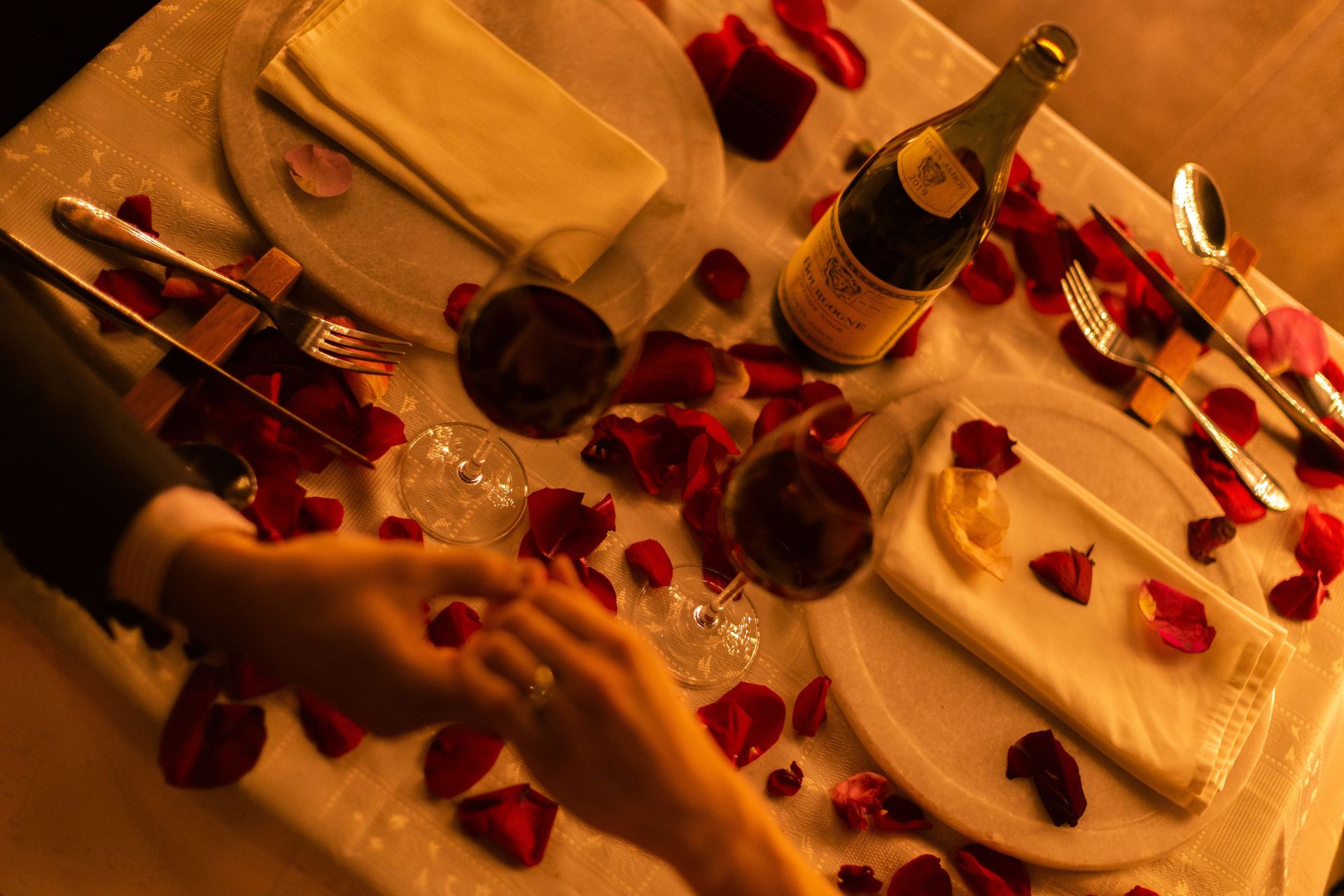 A candlelit table set for two, scattered with red rose petals, wine glasses and a bottle of Burgundy